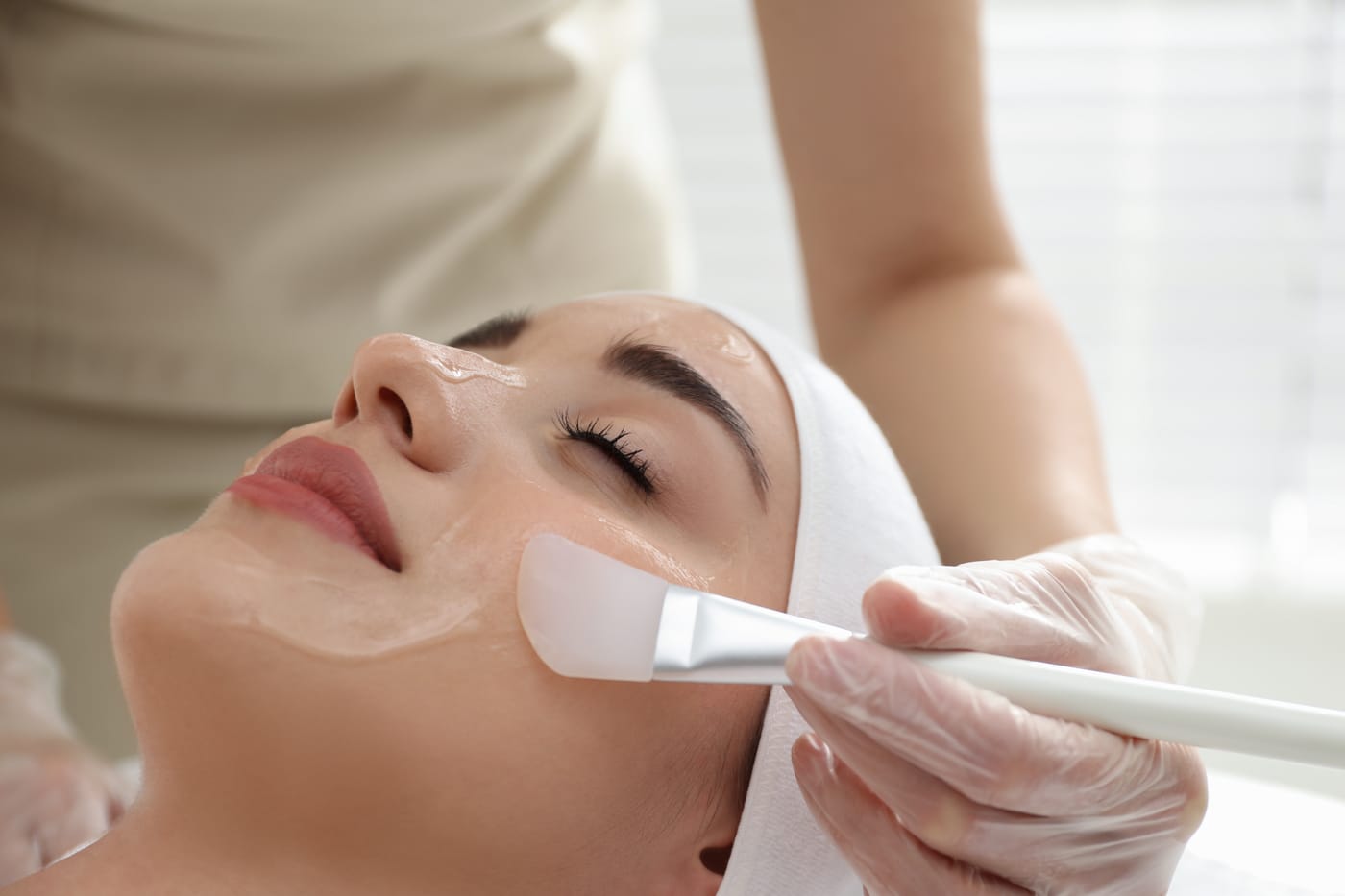 A gloved specialist brushes a chemical peel solution onto a relaxed woman's face