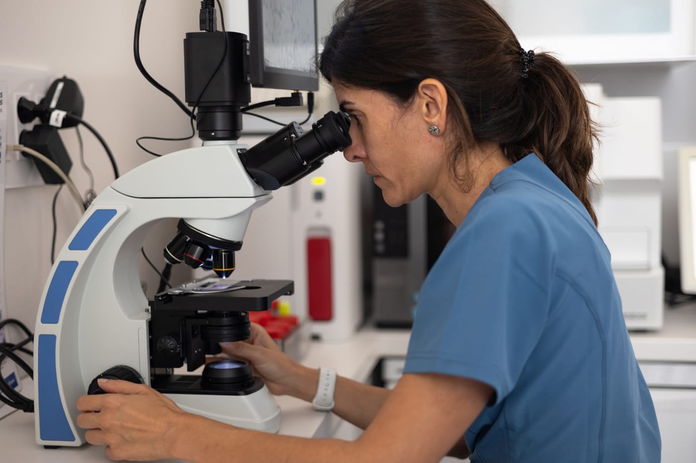 A clinician in scrubs examines a tissue sample under a microscope