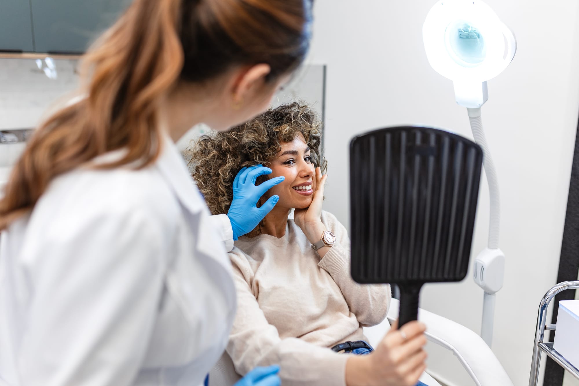 A dermatologist examines a smiling patient's face as she looks into a hand mirror